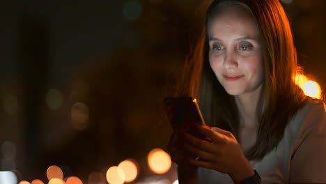 Una-Joven-En-La-Ciudad-Nocturna-Mira-La-Pantalla-Del-Teléfono-Inteligente-Y-Escribe-Un-Mensaje-De-Texto,-Se-Comunica-En-Las-Redes-Sociales,-Publica-Y-Comenta-Fotos